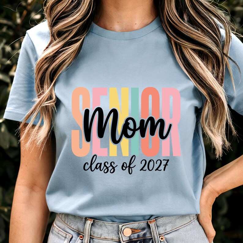 May include: A light blue short-sleeve t-shirt featuring the word SENIOR in large, colorful block letters behind the black cursive text Mom and the black script text class of 2027. The design is centered on the front of the shirt.