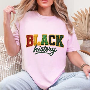 May include: A light pink t-shirt with the words "BLACK history" in a collegiate style. The word "BLACK" is in red, gold, green, and black. The word "history" is in a green script font.