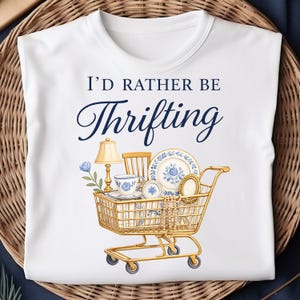 Op de afbeelding: Wit T-shirt met de tekst "I'd Rather Be Thrifting" in marineblauwe letters. Een gouden winkelwagenafbeelding is gevuld met vintage items, waaronder een lamp, stoel, theekopje en borden met blauwe bloemmotieven.