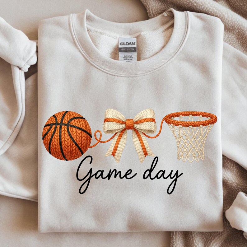 May include: Cream-colored sweatshirt featuring a basketball, bow, and hoop design. The basketball is orange with black lines, connected to a bow and a basketball hoop. The words "Game day" are written in a script font.