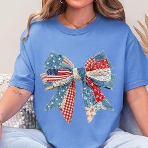 May include: Light blue t-shirt featuring a large bow design. The bow is made of patchwork fabric with denim, red gingham, floral, and lace details. An American flag is incorporated into the bow's design.