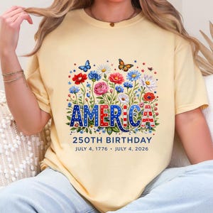 K&ouml;nnte beinhalten: Ein hellgelbes T-Shirt mit dem Wort "AMERICA" in Rot, Wei&szlig; und Blau, umgeben von einem Blumendesign mit Schmetterlingen. Der Text "250TH BIRTHDAY" und die Daten "July 4, 1776 &bull; July 4, 2026" stehen darunter.