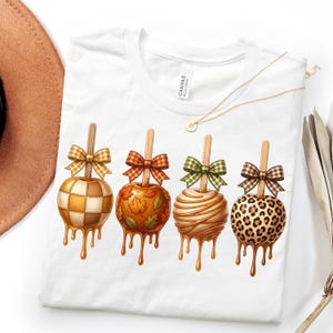 May include: A white t-shirt featuring a graphic of four caramel apples on sticks, each topped with a patterned bow. The apples display a checkered pattern, autumn leaves, drizzled caramel, and a leopard print design with dripping caramel.