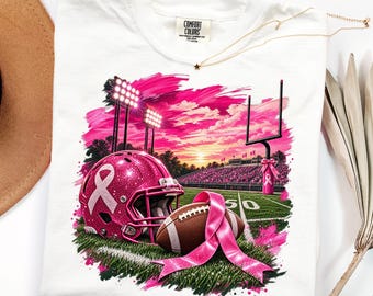 May include: A white t-shirt featuring a graphic print of a glittery pink football helmet with a breast cancer awareness ribbon, a brown football with a pink ribbon, a football field, stadium lights, and a sunset sky with pink clouds.