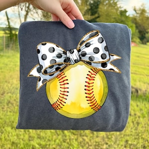 May include: A dark gray sweatshirt featuring a softball design. The design includes a yellow softball with red stitching, and a white bow with black polka dots and gold glitter trim. The sweatshirt is held up by a hand.