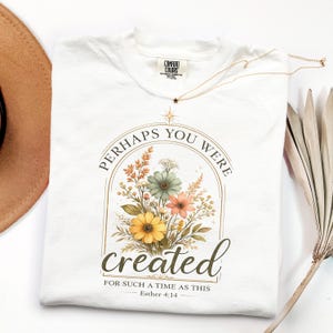 May include: A white t-shirt featuring a central graphic of a floral bouquet inside an arched frame. The text reads "PERHAPS YOU WERE created FOR SUCH A TIME AS THIS Esther 4:14" in a mix of serif and script fonts, accented by a small star.