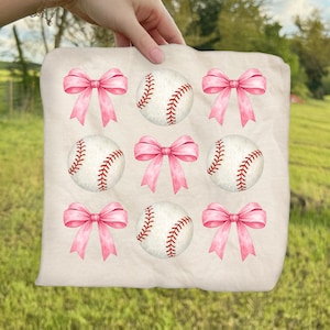 May include: A beige fabric with a pattern of pink bows and baseballs. The baseballs are white with red stitching. The bows are a solid pink color. The fabric is held up by a hand.