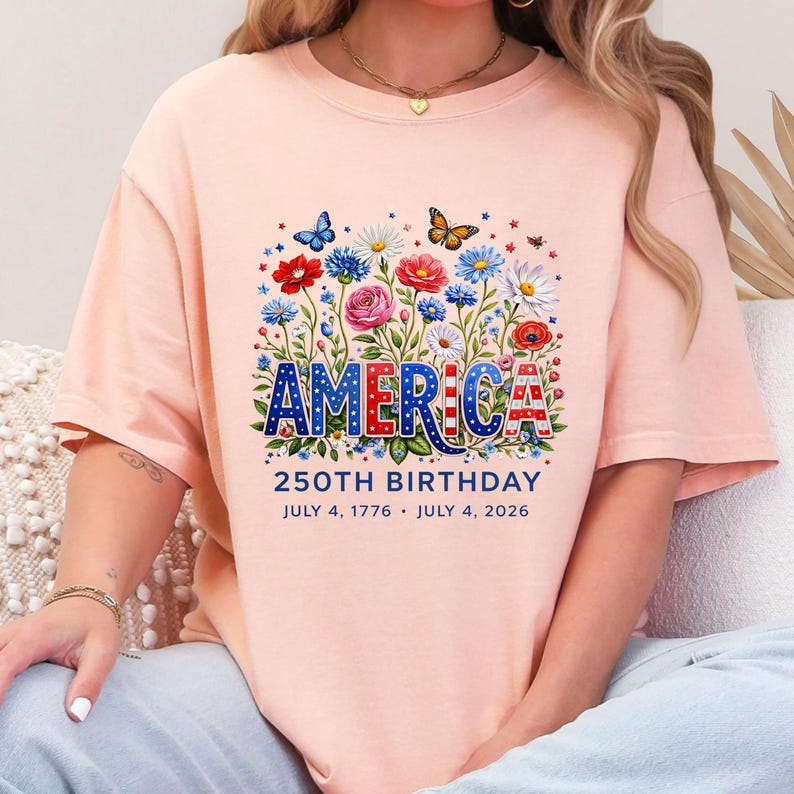K&ouml;nnte beinhalten: Pfirsichfarbenes T-Shirt mit floralem Design und dem Wort "AMERICA" in Rot, Wei&szlig; und Blau. Das Design zeigt Schmetterlinge und Blumen. Der Text "250TH BIRTHDAY JULY 4, 1776 &bull; JULY 4, 2026" steht darunter.