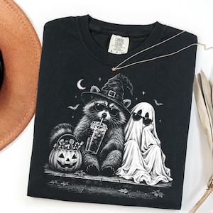 May include: A black t-shirt featuring a graphic of a raccoon wearing a witch hat and sunglasses, holding a drink, sitting next to a ghost wearing sunglasses. A jack-o'-lantern bucket filled with candy sits beside them under a crescent moon.