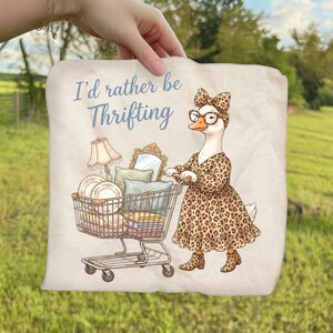 May include: A beige tote bag featuring a cartoon goose in a leopard print dress and glasses pushing a shopping cart filled with vintage items. The text "I'd rather be Thrifting" is written in blue script.