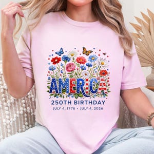 K&ouml;nnte beinhalten: Ein hellrosa T-Shirt mit dem Wort "AMERICA" in Rot, Wei&szlig; und Blau, umgeben von einem Blumendesign mit Schmetterlingen. Der Text "250TH BIRTHDAY" und die Daten "JULY 4, 1776 &bull; JULY 4, 2026" sind unter dem Wort "AMERICA" aufgedruckt.