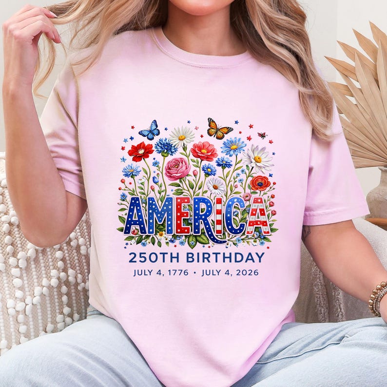 K&ouml;nnte beinhalten: Ein hellrosa T-Shirt mit dem Wort "AMERICA" in Rot, Wei&szlig; und Blau, umgeben von einem Blumendesign mit Schmetterlingen. Der Text "250TH BIRTHDAY" und die Daten "JULY 4, 1776 &bull; JULY 4, 2026" sind unter dem Wort "AMERICA" aufgedruckt.