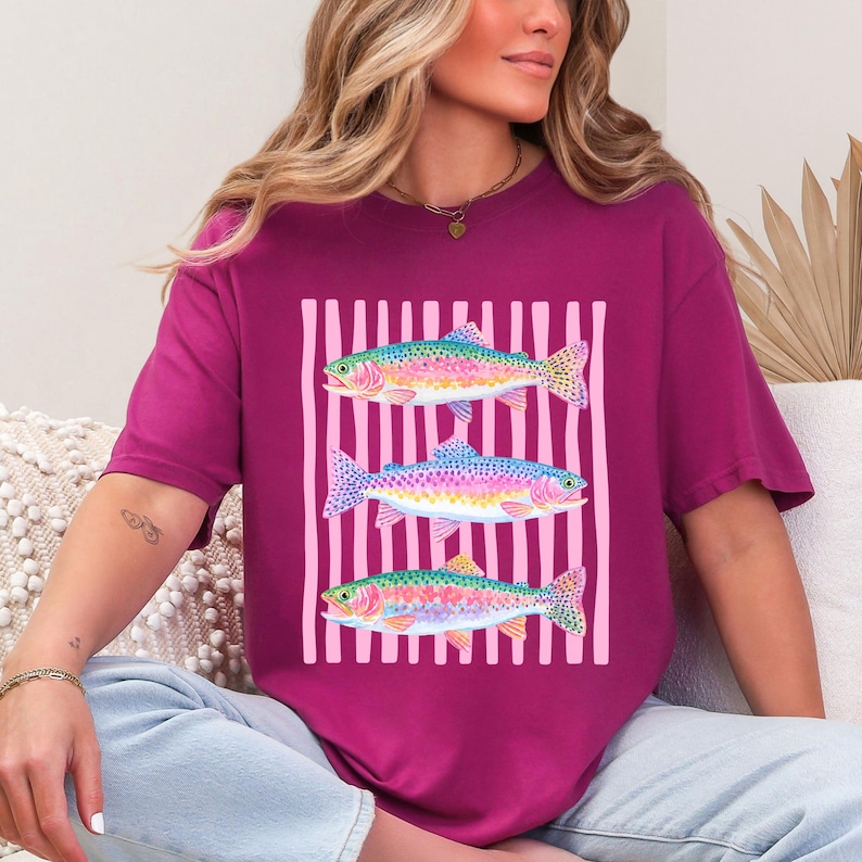 May include: A magenta t-shirt featuring a graphic of three rainbow trout. The fish are arranged vertically against a background of pink vertical stripes. The trout have colorful scales and fins.