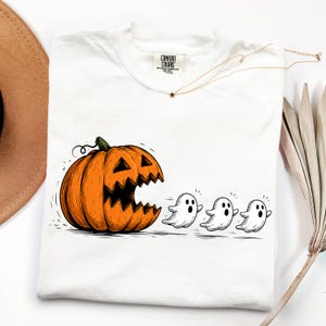 May include: A white t-shirt featuring a graphic of a large orange jack-o'-lantern with a wide, jagged mouth appearing to chase three small white ghosts. The shirt has a crew neckline and a Comfort Colors brand label printed on the inside collar.