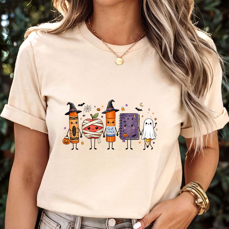 May include: A cream-colored t-shirt featuring a graphic print of five anthropomorphic school supplies dressed for Halloween, including a crayon, a mummy apple, a glue stick, a purple notebook, and a pencil dressed as a ghost.