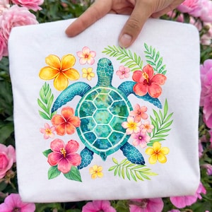 May include: White t-shirt featuring a watercolor design of a sea turtle surrounded by colorful tropical flowers and green leaves. The turtle has a blue shell and is centered on the shirt. The flowers are pink, orange, and yellow.