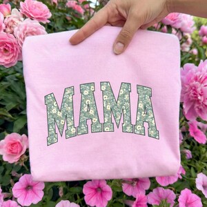 May include: A light pink sweatshirt with the word "MAMA" in large, floral-patterned letters. The letters are a muted green color with a white floral design. The sweatshirt is held up by a hand, with pink flowers in the background.