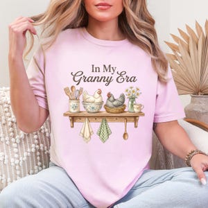 May include: A light pink t-shirt with the text "In My Granny Era" in brown script. The shirt features a watercolor illustration of a shelf with chickens, kitchen utensils, and flowers. Two dish towels and a wooden spoon hang below.