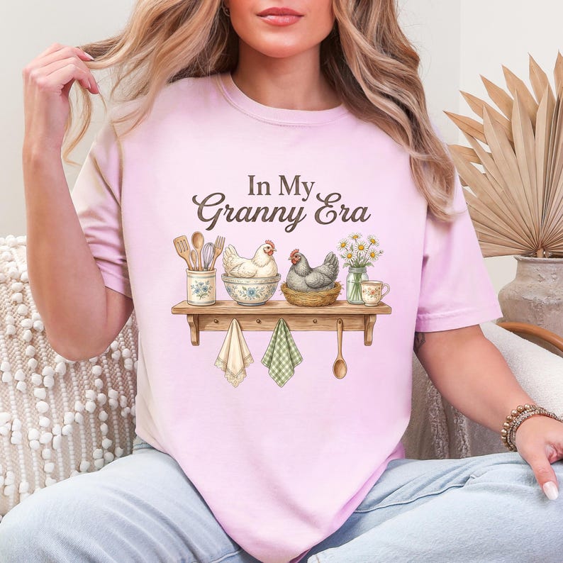 May include: A light pink t-shirt with the text "In My Granny Era" in brown script. The shirt features a watercolor illustration of a shelf with chickens, kitchen utensils, and flowers. Two dish towels and a wooden spoon hang below.