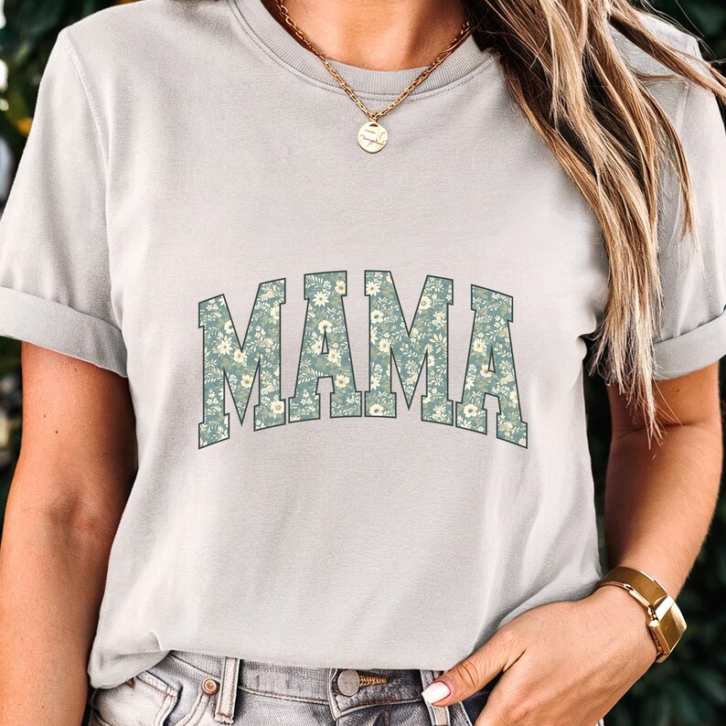 May include: A light gray t-shirt with the word "MAMA" in large, floral-patterned letters across the chest. The floral pattern features small white flowers on a green background. The shirt has short sleeves and a crew neck.