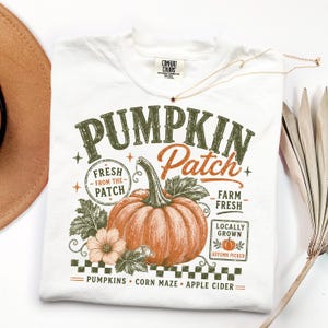 May include: A white t-shirt featuring a graphic of a large orange pumpkin with green leaves and a flower. The text reads PUMPKIN Patch, FRESH FROM THE PATCH, FARM FRESH, LOCALLY GROWN, AUTUMN PICKED, PUMPKINS, CORN MAZE, and APPLE CIDER in green and orange.