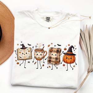 May include: A white t-shirt featuring a graphic print of four anthropomorphic characters: an open book wearing a witch hat, a steaming mug, a plaid blanket, and a pumpkin wearing a witch hat, all surrounded by small bats, spiders, and leaves.