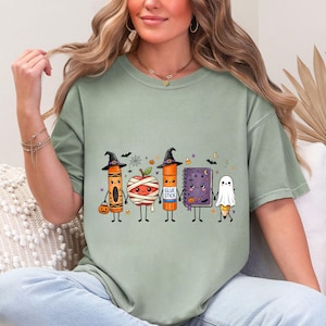 May include: A sage green t-shirt featuring a graphic of five cute, anthropomorphic school supplies dressed for Halloween: a crayon, a mummy apple, a glue stick labeled "GLUE STICK", a purple spiral notebook, and a pencil dressed as a ghost.