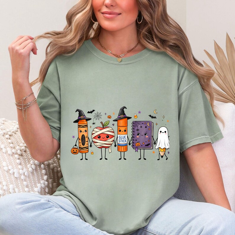 May include: A sage green t-shirt featuring a graphic of five cute, anthropomorphic school supplies dressed for Halloween: a crayon, a mummy apple, a glue stick labeled "GLUE STICK", a purple spiral notebook, and a pencil dressed as a ghost.