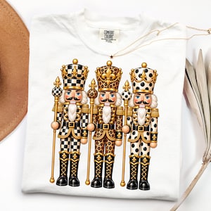 May include: A white t-shirt featuring a graphic print of three nutcracker figures standing side by side. Each nutcracker wears a crown and holds a gold staff, with outfits patterned in black and white checkers, leopard print, and black polka dots.
