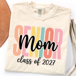 May include: A cream-colored t-shirt featuring the word SENIOR in large, vertical, multi-colored block letters in peach, pink, yellow, and teal. Overlaid in black script is the word Mom, with the text class of 2027 printed below in black.