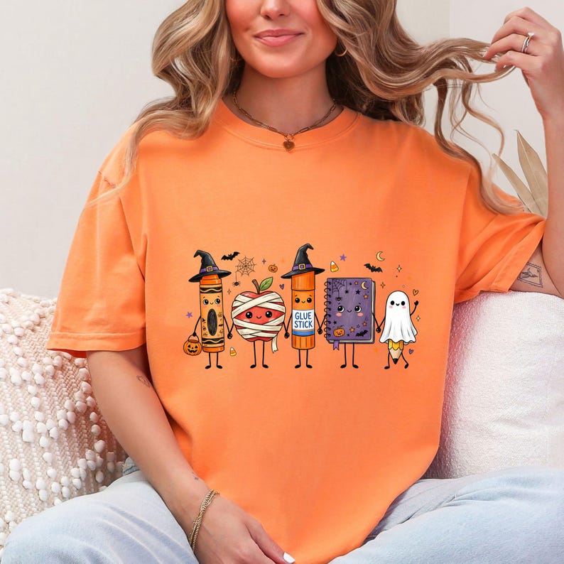 May include: An orange t-shirt featuring a graphic of five cute, anthropomorphic school supplies dressed for Halloween: a crayon in a witch hat, a mummy apple, a glue stick in a witch hat, a purple notebook, and a pencil dressed as a ghost.
