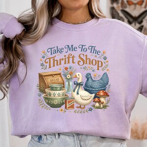 May include: A light purple sweatshirt with the text "Take Me To The Thrift Shop" and a graphic of a duck, chicken, bread box, bowls, and mushrooms. The graphic has a vintage, farmhouse aesthetic.