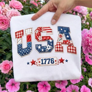 May include: White sweatshirt featuring the word "USA" in red, white, and blue patchwork lettering. The letters are decorated with floral and star patterns. Below the letters is the number "1776" in a rectangular design, with stars on either side.