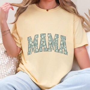 May include: A light yellow t-shirt with the word "MAMA" in large, block letters across the front. The letters are filled with a floral pattern in shades of blue and white. The shirt has short sleeves and a relaxed fit.