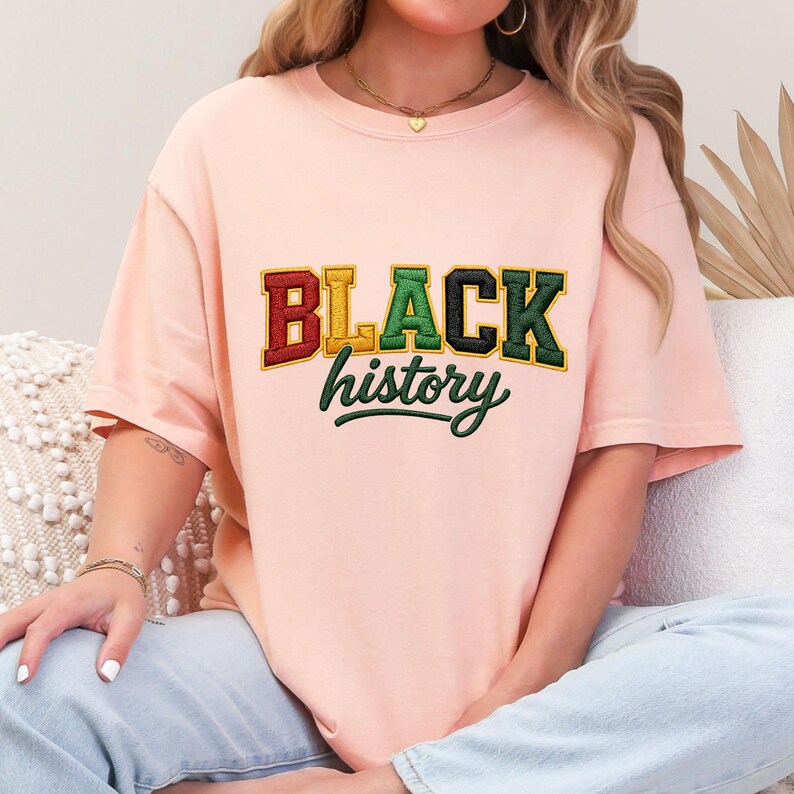 May include: A light peach-colored t-shirt with the words "BLACK history" embroidered in red, yellow, green, and black. The shirt has short sleeves and a crew neck. The text is in a collegiate style font.