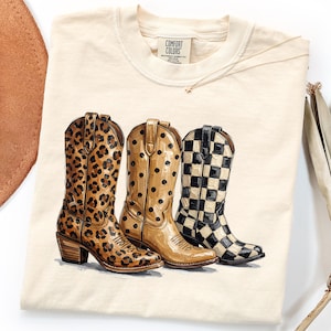 May include: Cream-colored Comfort Colors t-shirt featuring a graphic of three cowboy boots. The boots showcase distinct patterns: leopard print, tan with black polka dots, and a black and white checkerboard design. The shirt has a crew neck and a small tag.