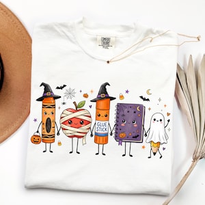 May include: A white t-shirt featuring a graphic of five cartoon characters: an orange crayon in a witch hat, a mummy-wrapped apple, an orange glue stick in a witch hat, a purple notebook, and a pencil dressed as a ghost, all holding hands.