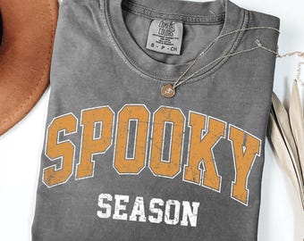 May include: A charcoal gray short-sleeve t-shirt with a distressed orange collegiate-style graphic that reads SPOOKY in an arch and SEASON in white block letters below. The shirt features a Comfort Colors brand label at the inner neckline.