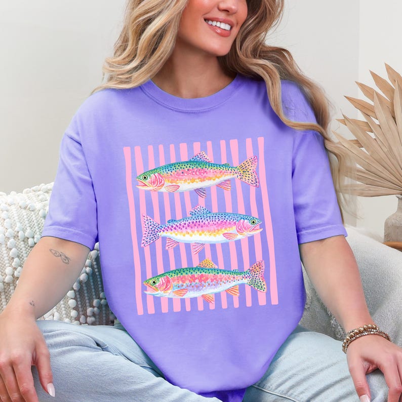 May include: A lilac-colored t-shirt featuring a graphic of three colorful fish. The fish are arranged vertically against a backdrop of pink vertical stripes. The fish have rainbow-colored scales and fins.