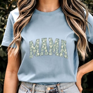 May include: A light blue t-shirt with the word "MAMA" in large, floral-patterned letters across the chest. The floral pattern features small white flowers on a green background. The shirt has short sleeves and a crew neck.