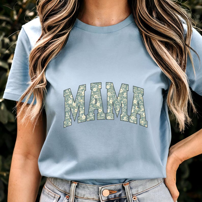 May include: A light blue t-shirt with the word "MAMA" in large, floral-patterned letters across the chest. The floral pattern features small white flowers on a green background. The shirt has short sleeves and a crew neck.