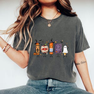 May include: A charcoal gray t-shirt featuring a graphic of five cute, anthropomorphic school supplies dressed for Halloween: a crayon in a witch hat, a mummy apple, a glue stick in a witch hat, a purple notebook, and a ghost-covered pencil.