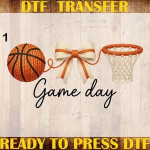 May include: A basketball, bow, and net graphic with the words "Game day." The design is in orange and white, with the text "DTF TRANSFER" and "READY TO PRESS DTF" in yellow. The background is a light wood grain.