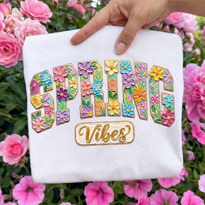 May include: White sweatshirt with the words "SPRING VIBES" in colorful, floral-filled letters. The letters are in a collegiate style, with a gold border around the words. The sweatshirt is held up by a hand, with pink flowers in the background.