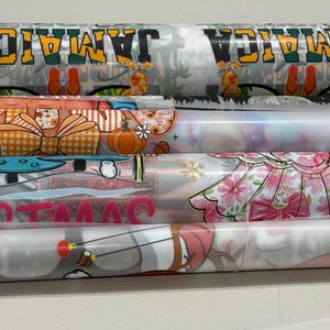 May include: Several rolls of patterned fabric are stacked horizontally. The top roll displays the word "JAMAICA" in black and yellow, with tropical imagery. Other rolls feature pumpkins, bows, floral designs, and the word "CHRISTMAS".