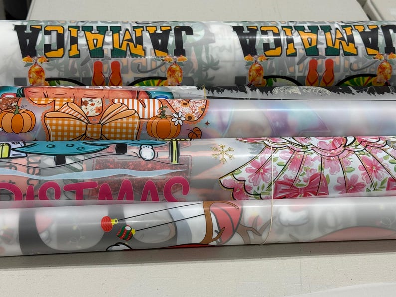 May include: Several rolls of patterned fabric are stacked horizontally. The top roll displays the word "JAMAICA" in black and yellow, with tropical imagery. Other rolls feature pumpkins, bows, floral designs, and the word "CHRISTMAS".