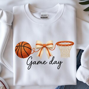May include: White sweatshirt with a basketball, bow, and hoop design. The basketball is orange with black lines, connected to a cream and orange bow, and a basketball hoop. The words "Game day" are written in black script.