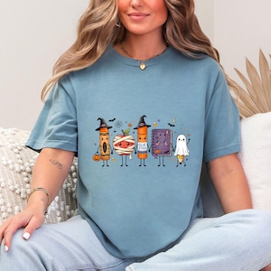 May include: A blue-gray crew neck t-shirt featuring a graphic of five anthropomorphic school supplies dressed for Halloween: a crayon, a mummy apple, a glue stick, a notebook, and a pencil ghost, all wearing witch hats or costumes.