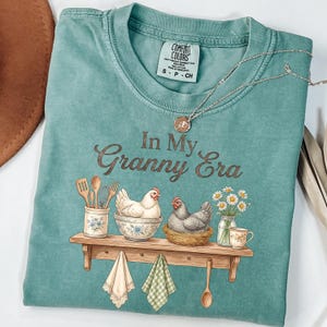 May include: A teal Comfort Colors t-shirt with the phrase "In My Granny Era" in brown script. The graphic features two chickens, kitchen utensils, and a shelf with a teacup and daisies. A silver necklace with a pendant is draped on the shirt.