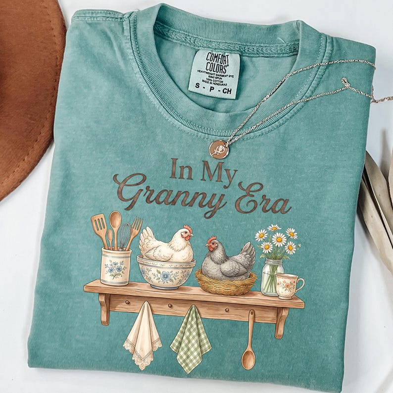 May include: A teal Comfort Colors t-shirt with the phrase "In My Granny Era" in brown script. The graphic features two chickens, kitchen utensils, and a shelf with a teacup and daisies. A silver necklace with a pendant is draped on the shirt.